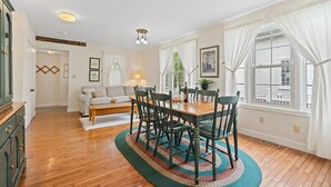 A dining area with a wooden table and chairs, a sofa, and a window with white curtains.