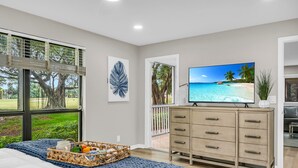 Ein Schlafzimmer mit großem Fenster, einem Bett mit blauer Decke, einem Schminktisch und einem Fernseher, der eine Strandansicht zeigt.