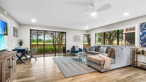 Ein geräumiges Wohnzimmer mit einer großen Couch, einem Couchtisch und einem Fernseher. Große Fenster bieten Blick auf Bäume und einen Balkon.
