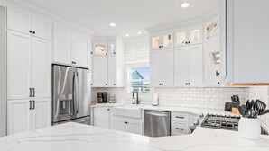 A modern kitchen with white cabinetry, a stainless steel refrigerator, a built-in oven, and a marble countertop. - Beach Retreat with Pool and Fire Pit (Holden Beach)
