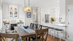 A modern kitchen with white cabinetry, a central island, and a dining area with wooden chairs and a table. - Beach Retreat with Pool and Fire Pit (Holden Beach)