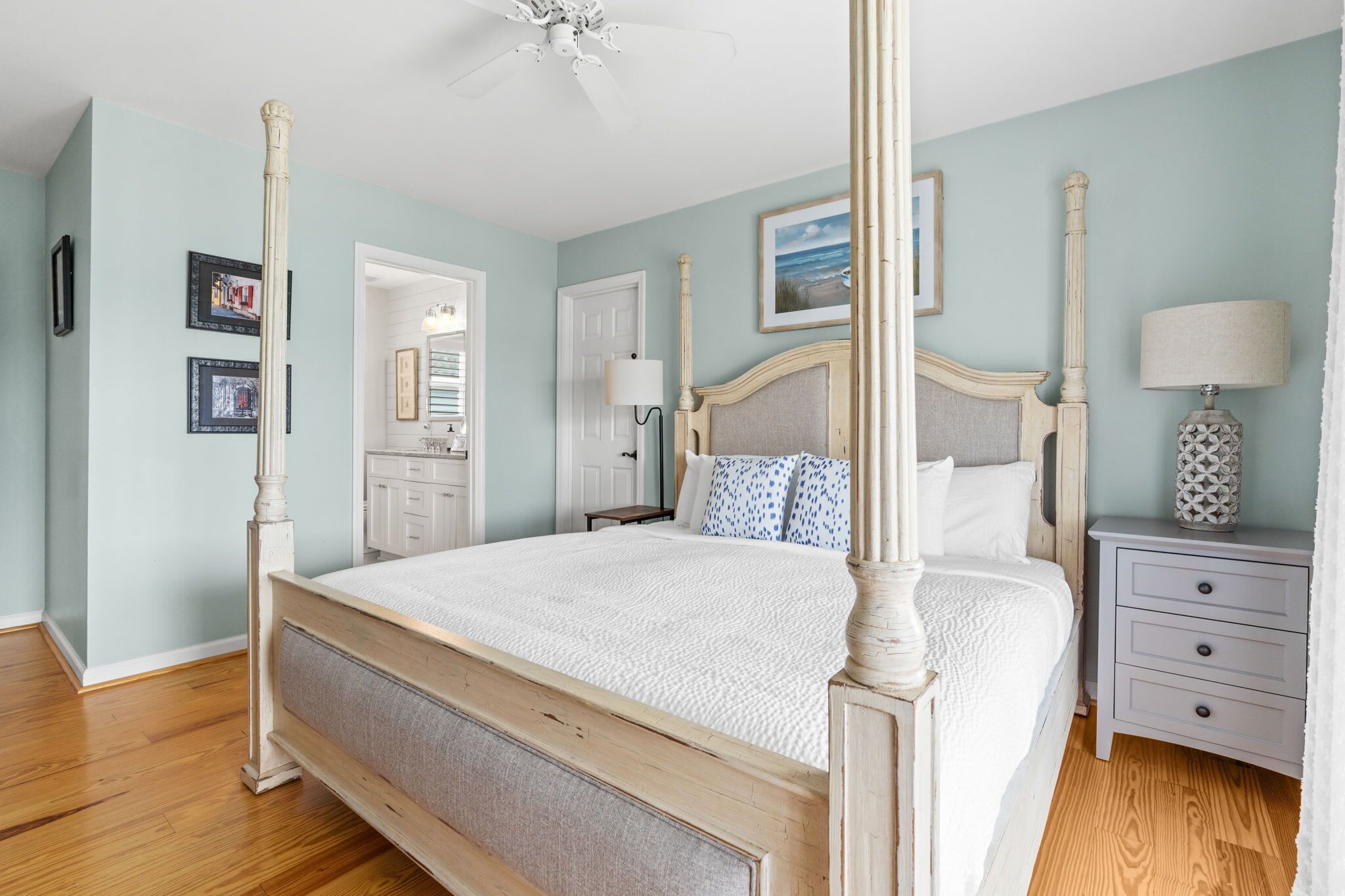 A four-poster bed with a wooden frame and a white bedspread, a nightstand with a lamp, and a door leading to another room.