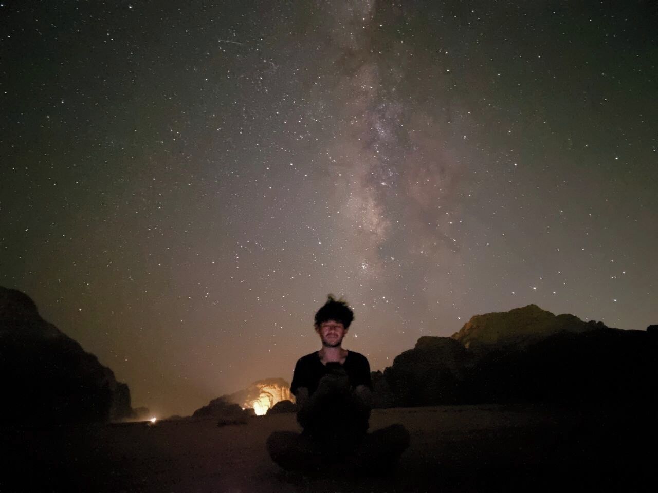 Eine Person sitzt auf einem Felsen unter einem sternenübersäten Himmel, wobei die Milchstraße deutlich sichtbar ist.