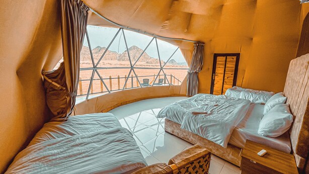 A room with two beds, a window with a view of a desert landscape, and a wooden door.