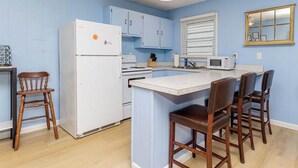 Eine Küche mit einem weißen Kühlschrank, Herd und einer Mikrowelle. Es gibt Holzstühle und eine kleine Kochinsel mit weißer Arbeitsplatte.