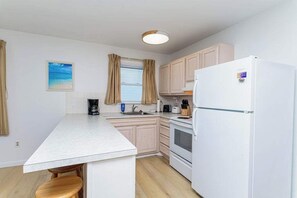 Eine kleine Küche mit weißer Arbeitsplatte, Holzschränken, einem Kühlschrank und einem Fenster mit Vorhängen.