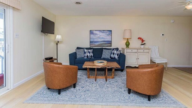 A living room with a blue sofa, two brown armchairs, a wooden coffee table, a TV mounted on the wall, and a painting above the sofa. - Midtown OC Unit 4/ Oceanblock / Sleeps 8 (Ocean City)