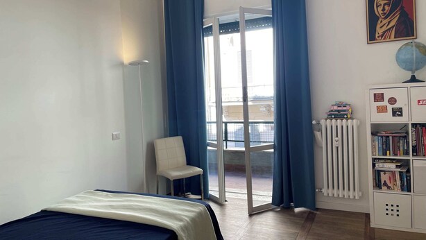 Una camera da letto con un letto, una sedia, un termosifone, una libreria e vista attraverso una porta in vetro.