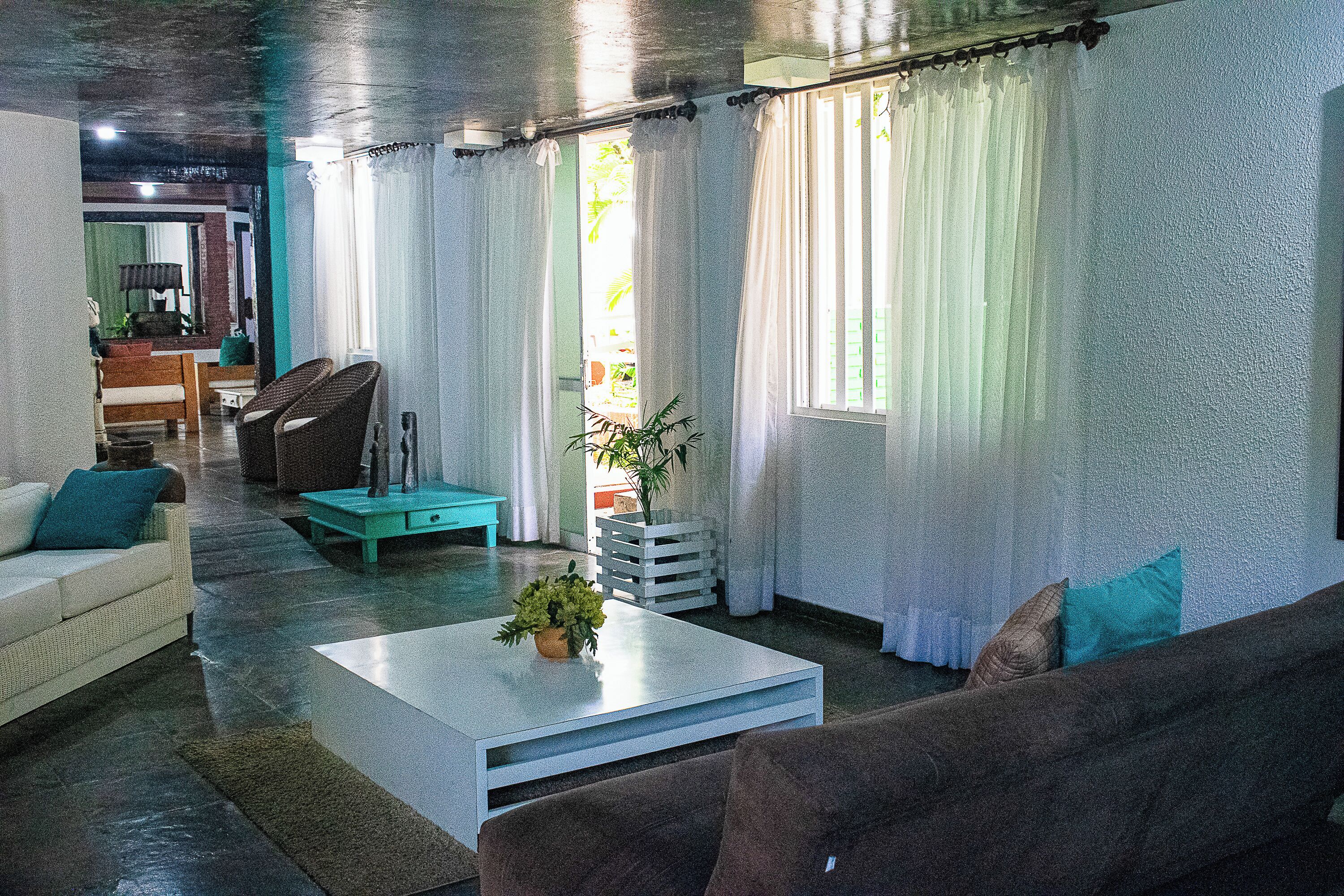 A living room with a white coffee table, a brown sofa, and a teal armchair. There is a plant on the coffee table and a window with curtains.