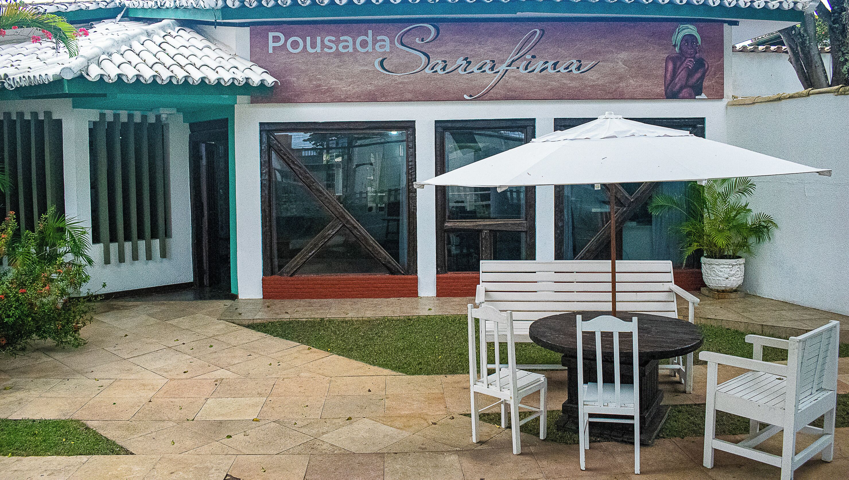 A patio area with a white bench, a table, and an umbrella. There is a sign above the entrance that reads "Pousada Sarafina."