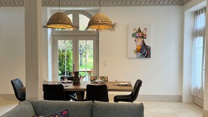 A modern living room with a grey sectional sofa, a dining area with a table and chairs, and a large window with a view of greenery.