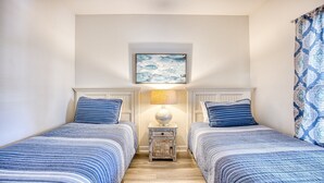 A bedroom with two beds, a nightstand with a lamp, a framed picture on the wall, and a patterned curtain. - 1521 Ironwood - Barefoot Resort Rentals (North Myrtle Beach)