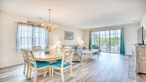 A spacious dining area with a wooden table and chairs, a chandelier, and a large window with curtains. - 1521 Ironwood - Barefoot Resort Rentals (North Myrtle Beach)