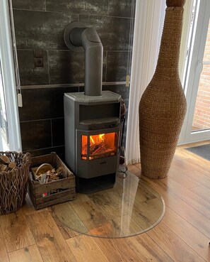 A wood-burning stove with a visible fire, placed in a room with a tiled wall and a large woven vase.