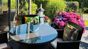 Um pátio ao ar livre com uma mesa de vidro redonda, cadeiras de vime, um candeeiro e plantas em vasos.