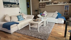 Uma sala de estar moderna com um sofá branco, uma mesa de centro e um tapete felpudo cinza. O chão é de madeira e a parede de tijolos tem prateleiras com objetos decorativos.