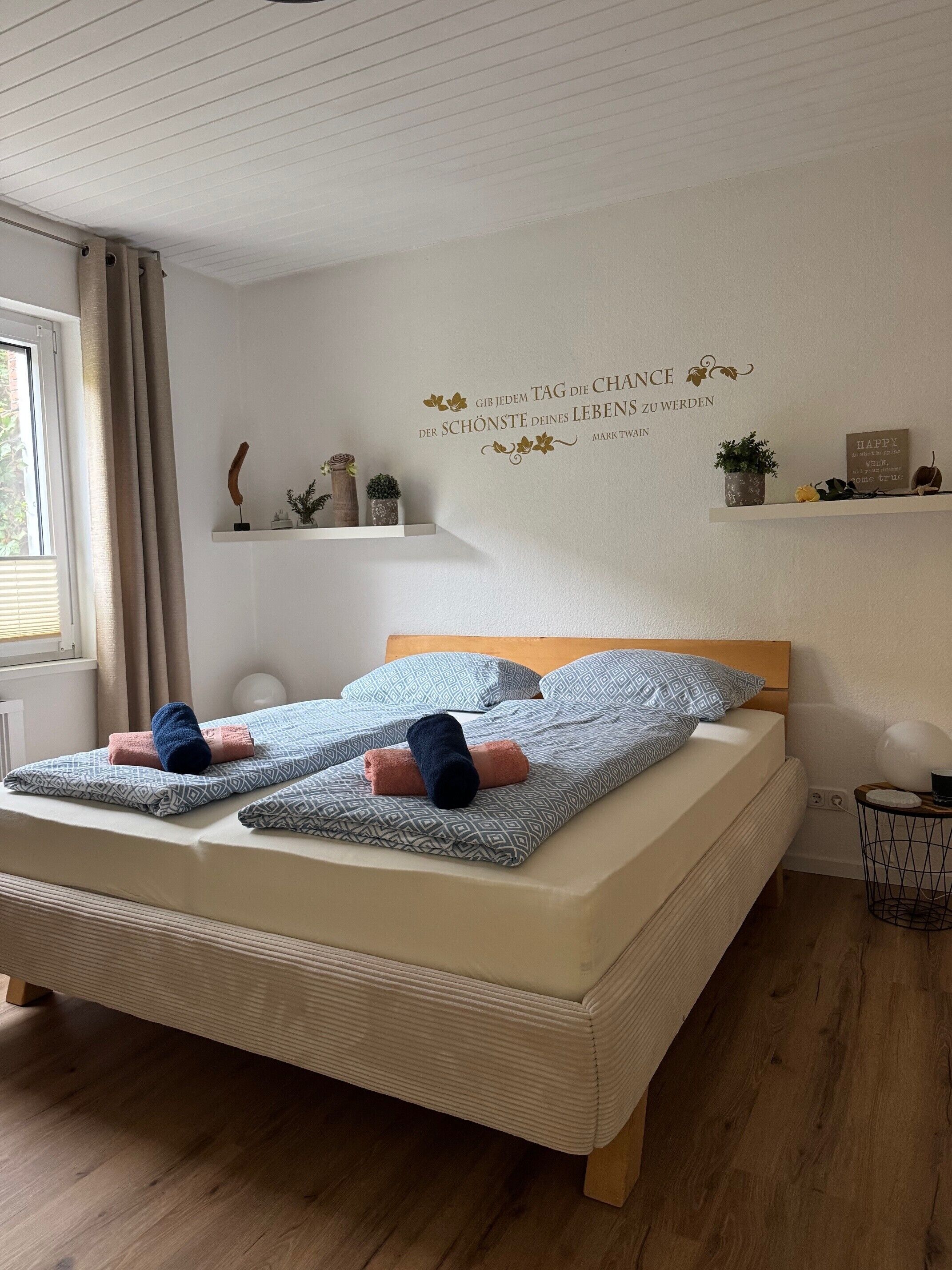 Une chambre à coucher avec un lit, des oreillers, une table de chevet, un élément de décoration murale et une fenêtre avec des rideaux.
