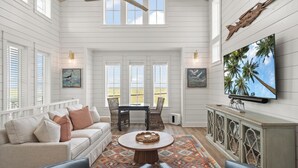 A spacious room with a high ceiling, white wooden walls, a large window, a ceiling fan, a white sofa, a dining table with chairs, a TV, and a decorative cabinet. - Home w/ Bay Views, Resort Amenities & Beach Access @Cinnamon Shore North! (Port Aransas)