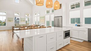 Una cocina amplia con una isla grande, armarios blancos y un refrigerador de acero inoxidable. Hay lámparas de mimbre colgantes y ventanas grandes que dejan entrar luz natural.