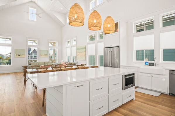 Una cocina amplia con una isla grande, armarios blancos y un refrigerador de acero inoxidable. Hay lámparas de mimbre colgantes y ventanas grandes que dejan entrar luz natural.