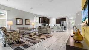 A modern living room with a sofa, armchair, and coffee table, a kitchen with dark cabinets and stainless steel appliances, and a large TV on the wall. - Magical 7BR Disney Retreat | Pool, Spa, Game Room & Theater! (Kissimmee)