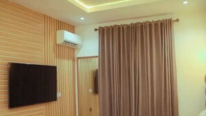 A hotel room with a flat-screen TV mounted on the wall, a bed with white bedding, a window with brown curtains, and a ceiling with recessed lighting.