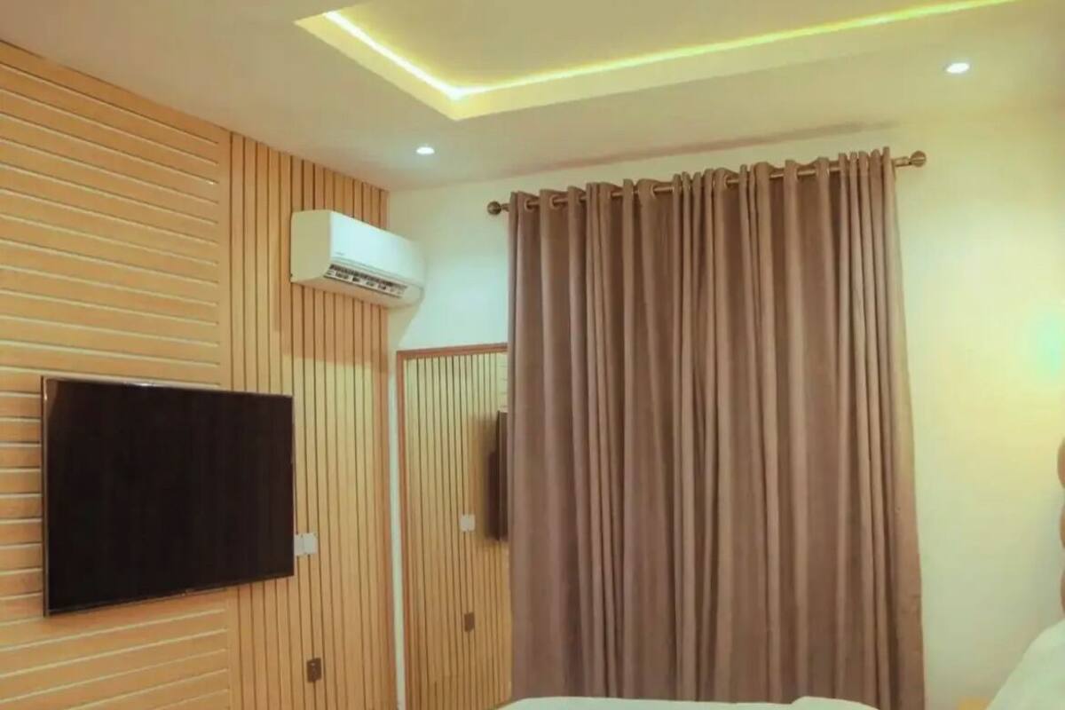 A hotel room with a flat-screen TV mounted on the wall, a bed with white bedding, a window with brown curtains, and a ceiling with recessed lighting.