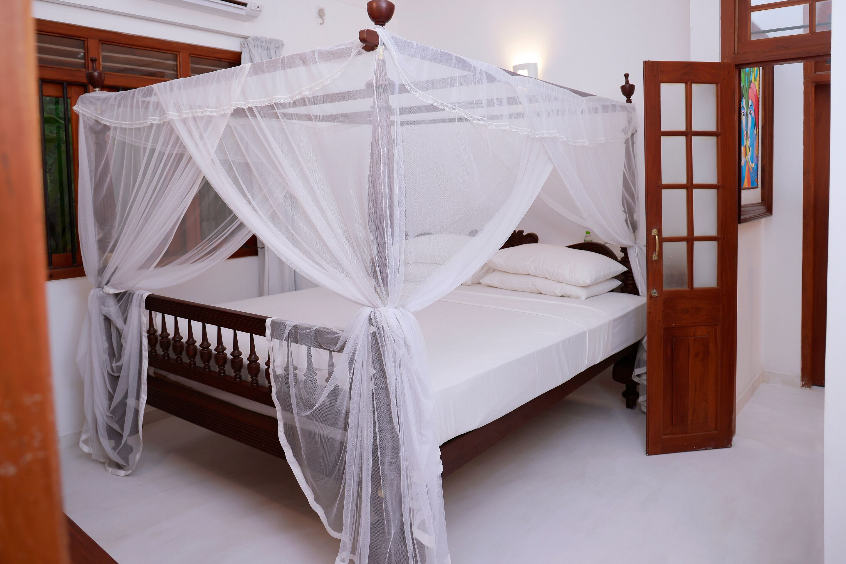 Una cama con dosel blanco y cabecera de madera, una puerta de madera con paneles de vidrio y un cuadro en la pared.