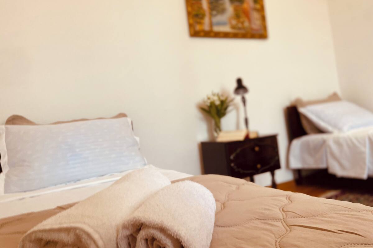 A neatly made bed with a folded towel, a bedside table with a lamp and a vase of flowers, and a small nightstand with a book.