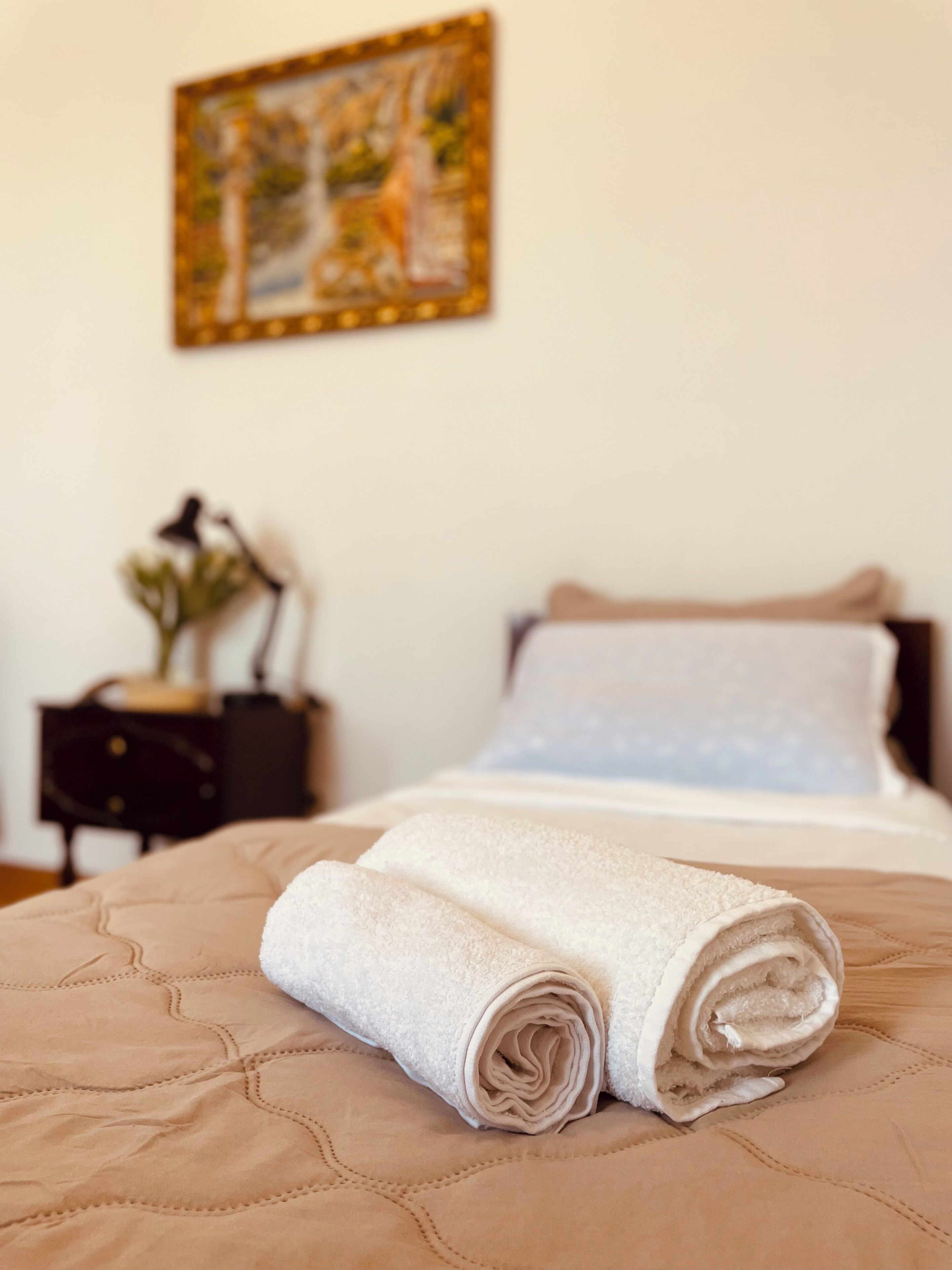 Cama bem arrumada com um edredom bege e duas toalhas brancas enroladas, mesa de cabeceira com uma luminária e um vaso, e um quadro na parede.