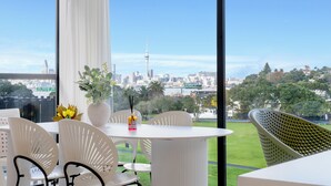 Un comedor moderno con sillas blancas y una mesa, ventanas amplias con vista a la ciudad, y un jarrón con flores.