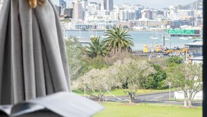 Vista desde una ventana con cortina, mostrando un paisaje urbano con una torre destacada, un puerto deportivo y un campo verde.