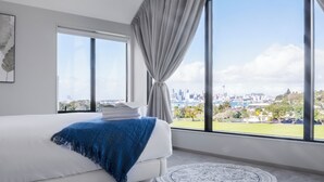 Un dormitorio moderno con un gran ventanal, una cama blanca y vistas a un horizonte urbano.