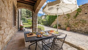 An outdoor dining area with stone walls, a table set with food and drinks, and an umbrella providing shade. - Caravaggio, Galenda, Gaiole in Chianti, Siena and Chianti (Gaiole in Chianti)