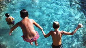 Ein klares Schwimmbecken mit sichtbaren Felsen unter Wasser und einem Kind, das die Hand eines Erwachsenen hält.
