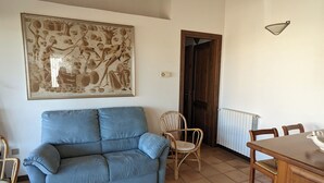 Ein Wohnzimmer mit einem blauen Sofa, einem Korbstuhl, einem Esstisch mit Stühlen und einem großen gerahmten Bild an der Wand.