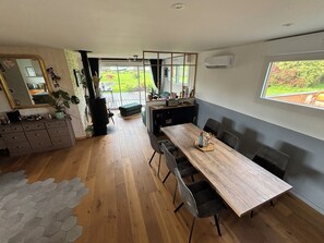 A modern kitchen with a large dining table, wooden flooring, and a view of the garden through the glass door. - Maison en Bois , Ty Kérou (Mahalon)