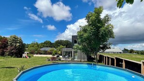 Una piscina con un deck circundante, una pequeña estructura de juegos y una casa moderna al fondo.