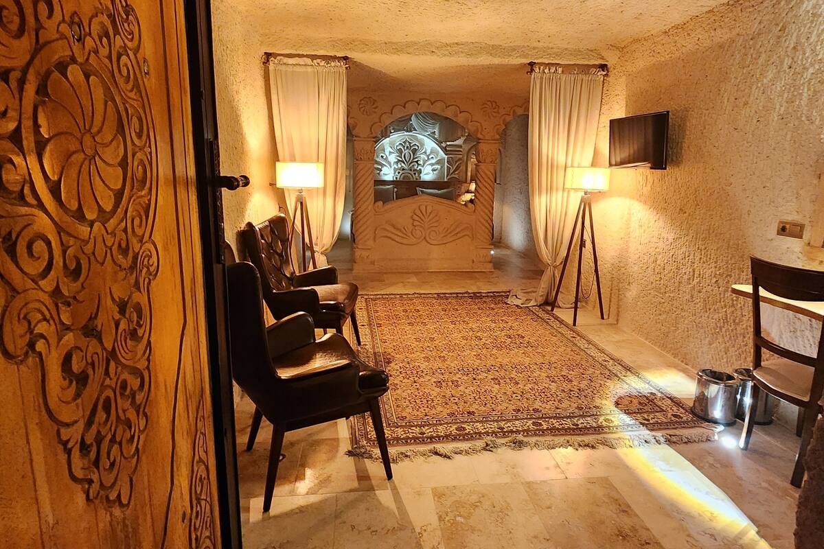 A room with a patterned carpet, a bed with a headboard, two standing lamps, a television on the wall, and a wooden door with intricate carvings.