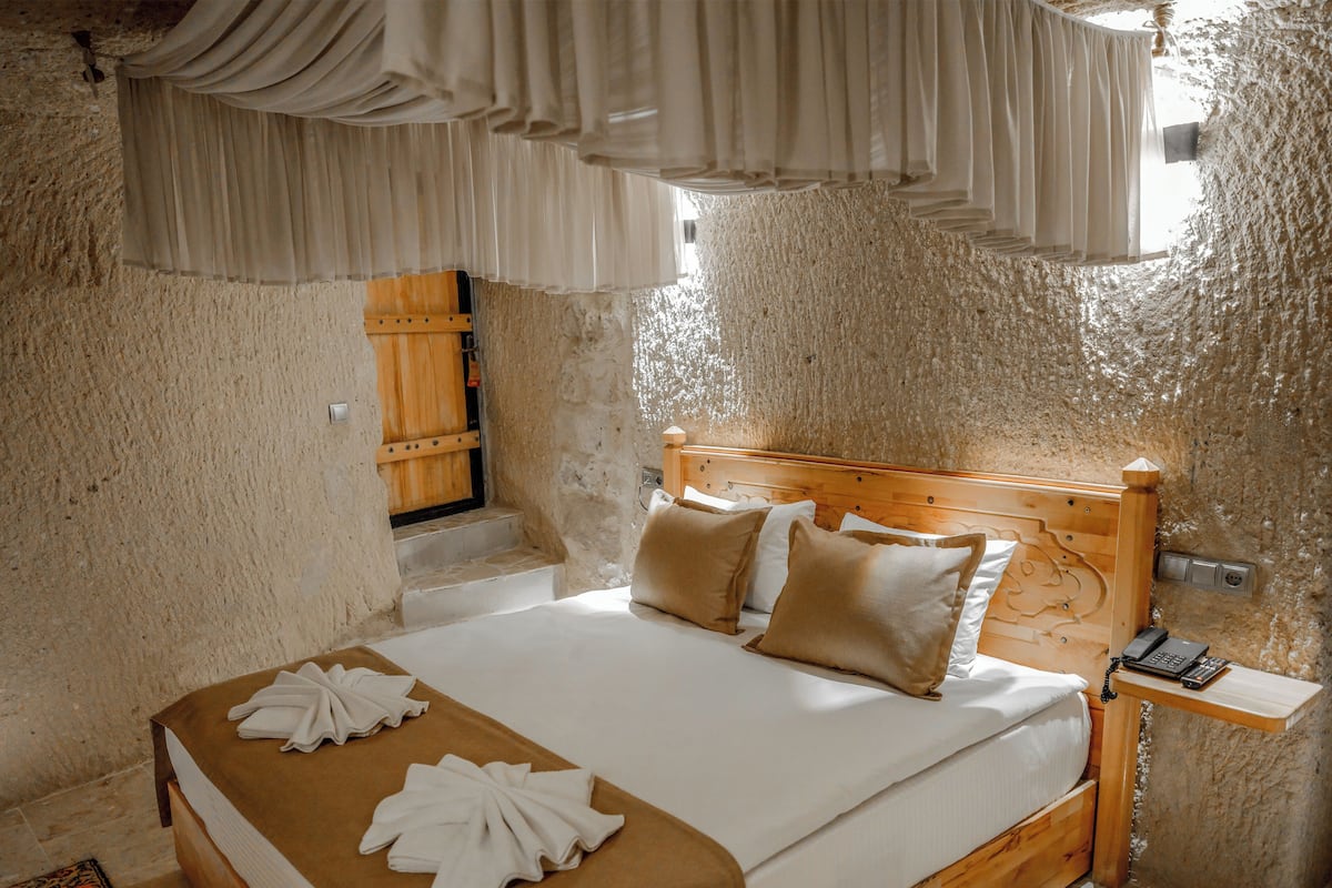 A bedroom with a wooden headboard, a bed with white linens and beige pillows, a wooden nightstand with a phone, and a window with curtains.