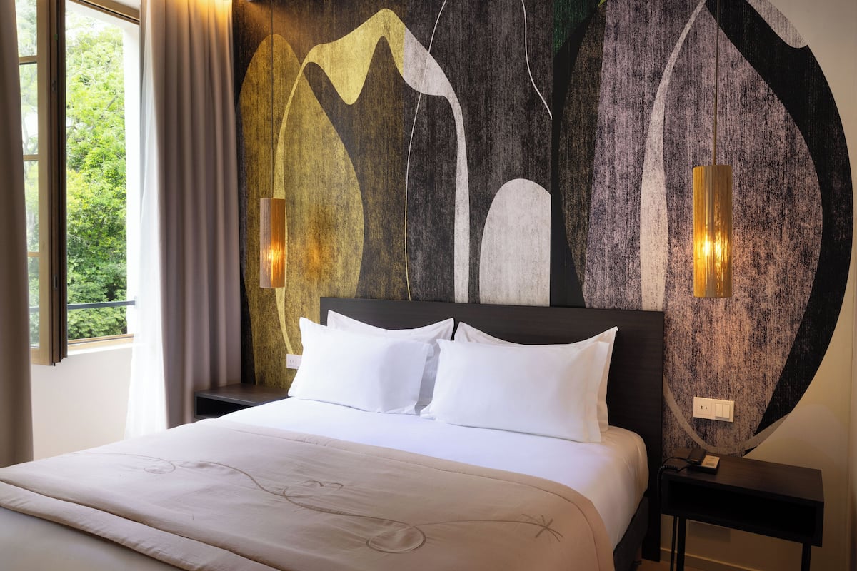 A hotel room with a large bed, white bedding, a dark headboard, and a wall mural. There are two wall-mounted lamps and a window with curtains.