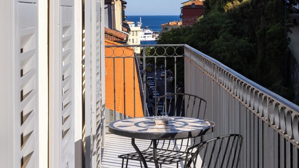 A balcony with a round table and chairs, overlooking a street with buildings and a view of the sea. - Palais Ségurane Nice Boutique Hôtel (Nice)