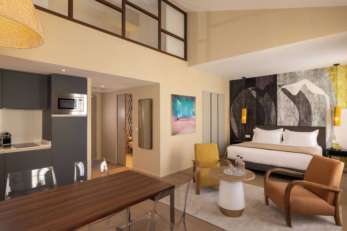 A modern hotel room with a dining area, a bed with a large painting, and a view of the city.