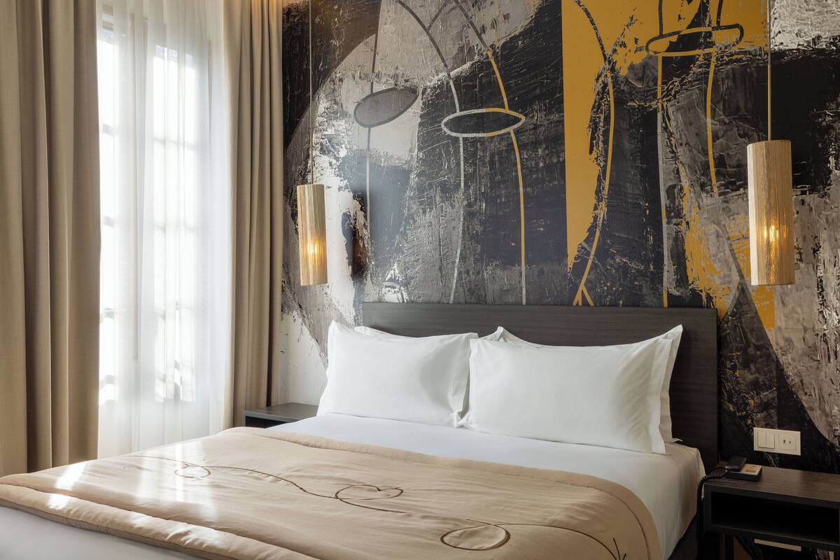 A hotel room with a large bed, a headboard with a light fixture, and a wall with abstract art.