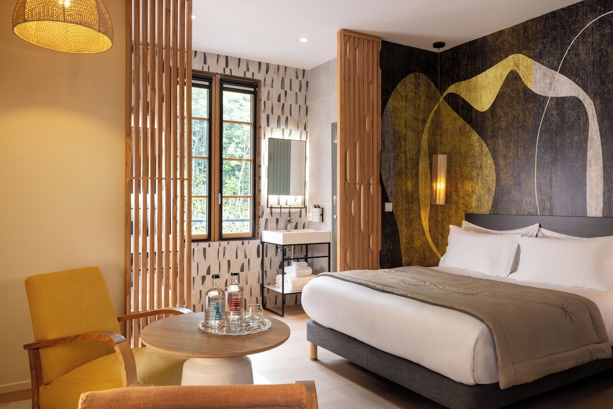 Habitación de hotel moderna con una cama grande, una mesa redonda para comer, un sillón amarillo, un separador de madera y vistas al exterior.