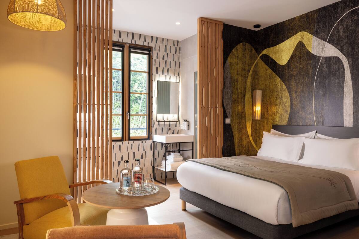 A modern hotel room with a large bed, a round dining table, a yellow armchair, a wooden partition, and a view of the outdoors.