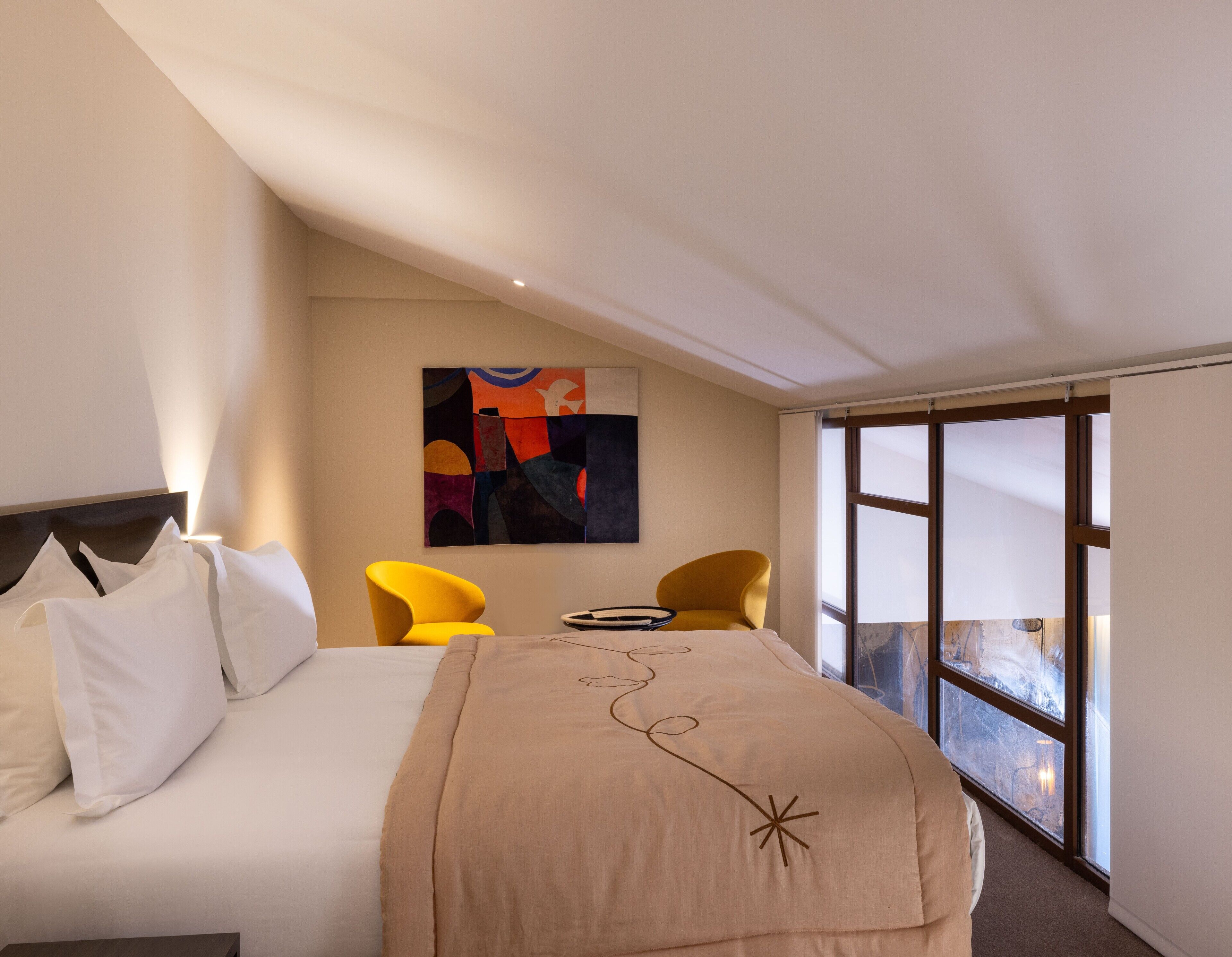 Et hotelværelse med en seng, to gule stole, et bord, et billede på væggen og et stort vindue.