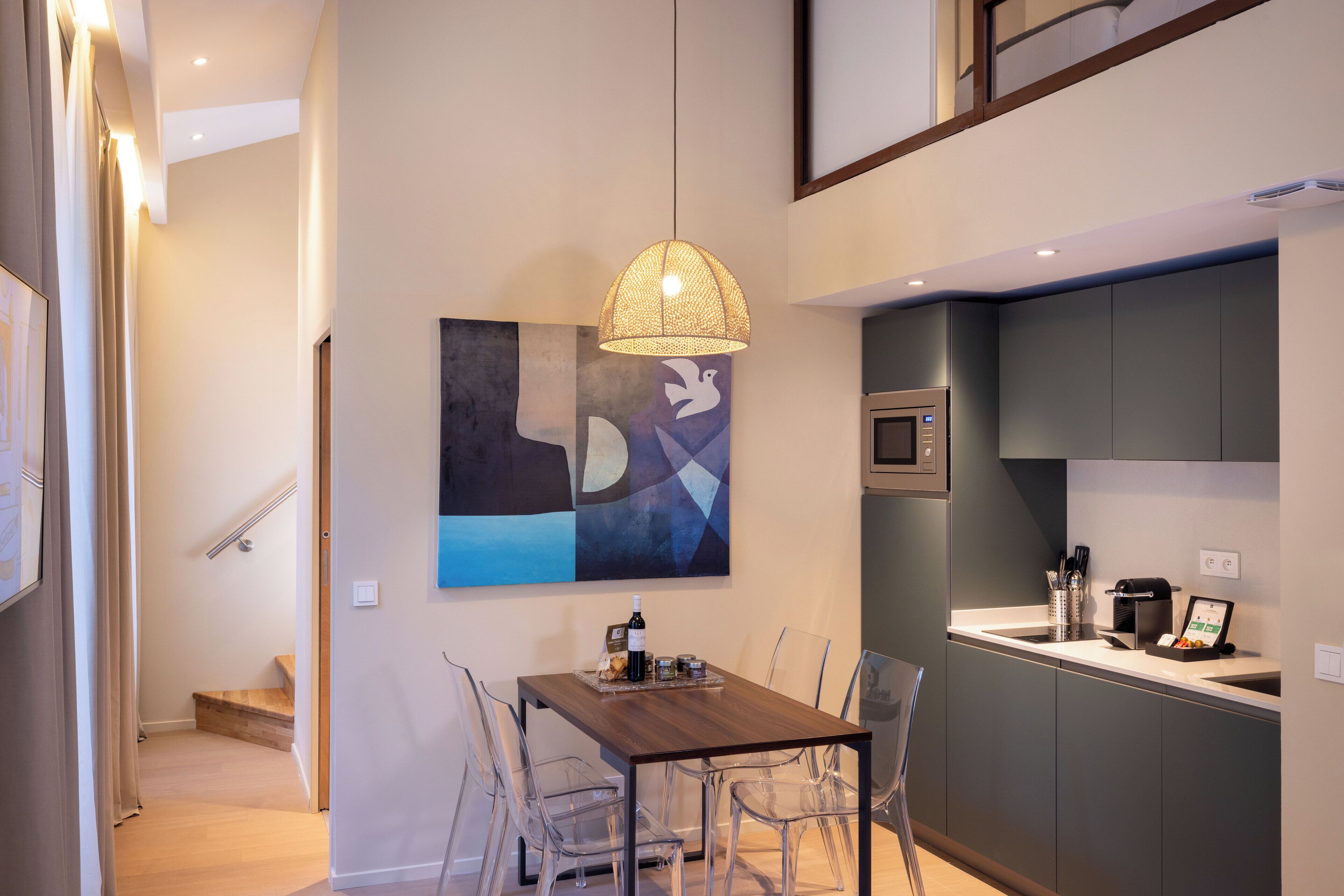 A modern kitchen with a dining area, featuring a wooden table and clear chairs, a hanging light fixture, and a painting on the wall.