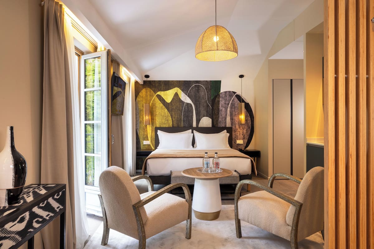 A modern hotel room with a large bed, two chairs, a round table, and a large abstract painting on the wall.