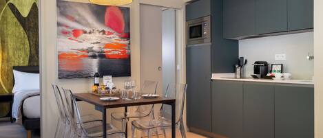 A modern kitchen with grey cabinets, a microwave, and a coffee machine. There is a dining table with clear chairs and a painting on the wall.